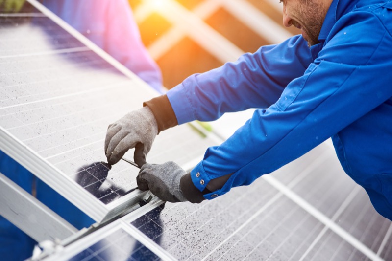 Smiling male technician in blue suit installing photovoltaic blue solar modules with screw