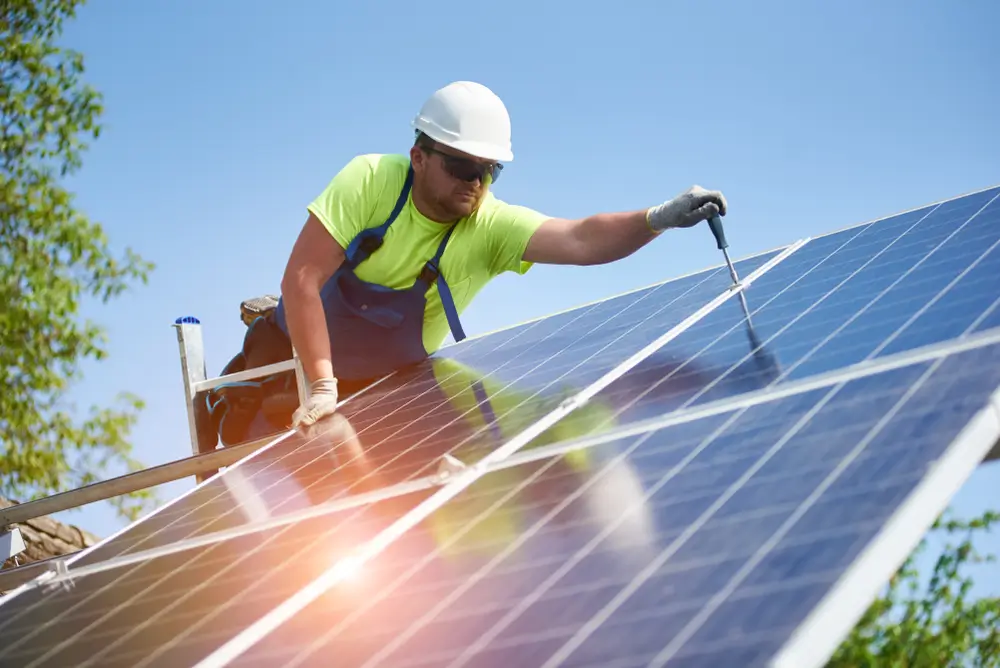 Technician connecting solar photo voltaic panel to metal platform using screwdriver standing on ladder on bright blue sky copy space background. Stand-alone solar panel system installation concept.
